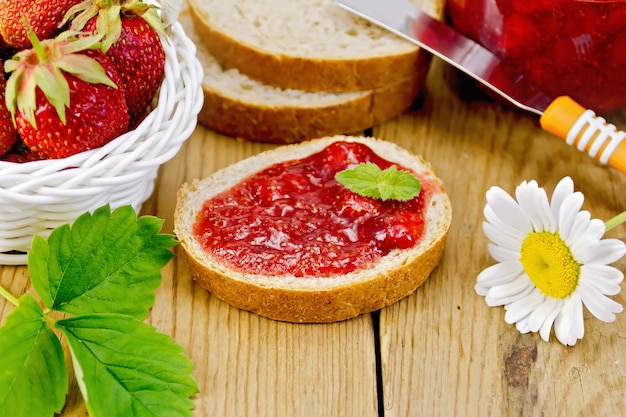 Pan con mermelada de fresa, un tarro de mermelada, cuchillo, fresas en una cesta de mimbre, manzanilla sobre un fondo de tablas de madera