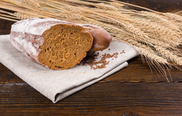 Pan de pan de centeno de panadería cortado por la mitad sobre un paño marrón junto a semillas y tallos de trigo secos sobre mesa de madera