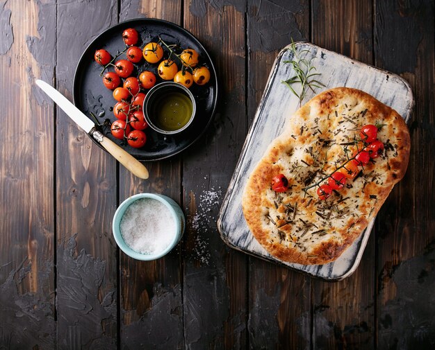 Pan plano de focaccia de romero casero servido con tomates al horno, aceite de oliva y romero fresco, sal marina y cuchillo sobre un fondo de madera. Vista superior