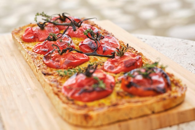 Pan plano de forma rectangular con rebanadas de tomate y ramas de tomillo en una tabla de cortar de madera sobre un fondo borroso