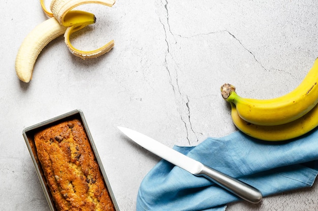 Un pan de plátano con trocitos de chocolate, algunos plátanos y una servilleta azul, sobre fondo gris.