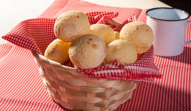 Pan de queso, canasta forrada con tela roja y blanca llena de pan de queso sobre una toalla a cuadros y una taza blanca.