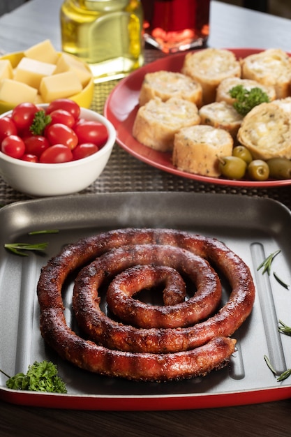 Foto pan de salchicha y ajo en un plato rojo sobre la mesa de barbacoa con aperitivos, queso, romero, aceitunas y tomates cherry.