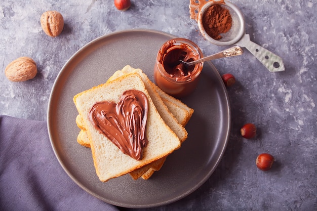 Pan tostado con mantequilla de crema de chocolate en forma de corazón, tarro de crema de chocolate