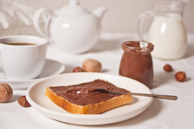 Pan tostado con mantequilla de crema de chocolate, tarro de crema de chocolate, cucharada de té, tarro de leche, tetera en el blanco.