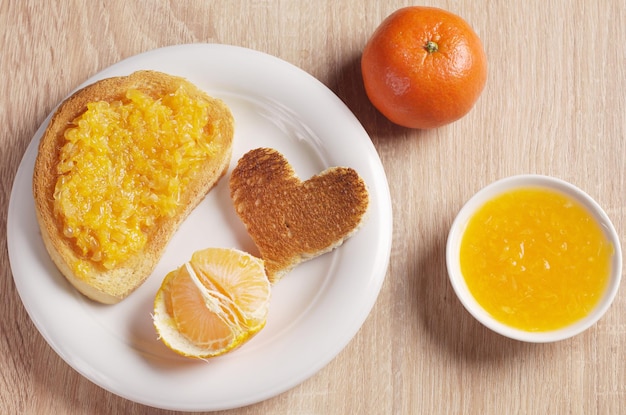 Pan tostado con mermelada de mandarina, rebanada en forma de corazón en un plato y fruta mandarina en la mesa de madera, vista superior