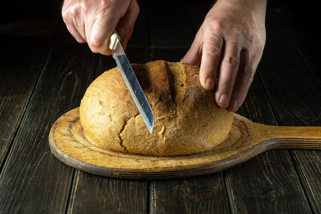 Pan de trigo en una tabla de cortar de madera recién salido del horno Chef mano con un cuchillo para cortar pan integral fresco