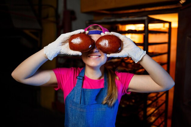 Una panadera profesional sostiene bollos frescos en sus manos y escucha su olor en la panadería. Lleva un vestido de mezclilla y una gorra. Elaboración de productos de panadería. Rejilla con bollería crujiente caliente.