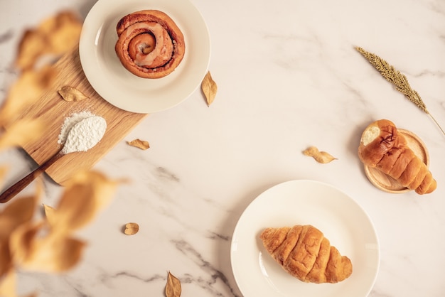 Panadería fresca, bollo de canela y croissant fresco en placa con cuchara de madera llena de harina sobre tabla de madera. Sabroso postre delicioso en la superficie de mármol blanco. Desayuno francés Vista superior