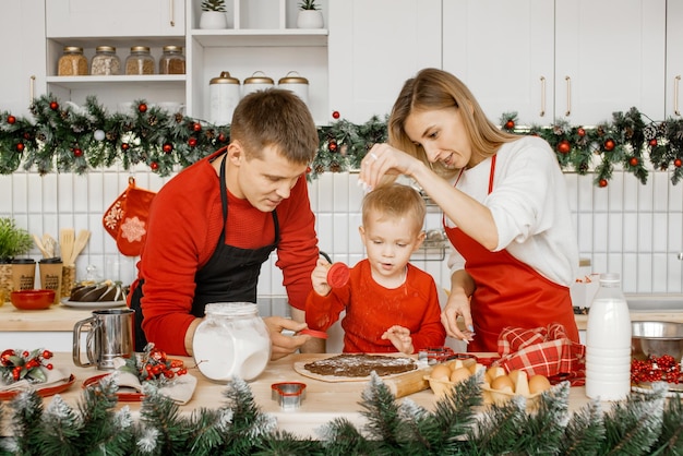 Panadería navideña. Familia junta haciendo pan de jengibre en la cocina festiva, padre e hijo preparándose para cortar galletas de masa de pan de jengibre mientras la madre esparce harina sobre la masa.