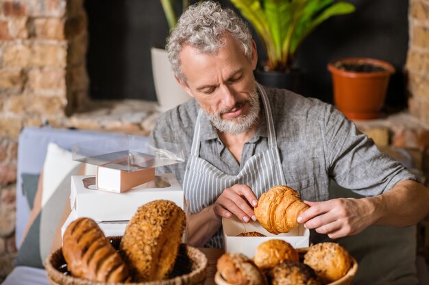 Panadería, pequeña empresa. Hombre barbudo canoso atento concentrado cuidadosamente embalaje croissant en caja sentado a la mesa en la pastelería