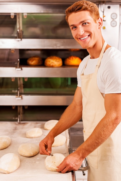 Foto panadero confiado en el trabajo. hombre joven confiado en delantal trabajando con masa y sonriendo mientras está de pie contra el horno con pan en él