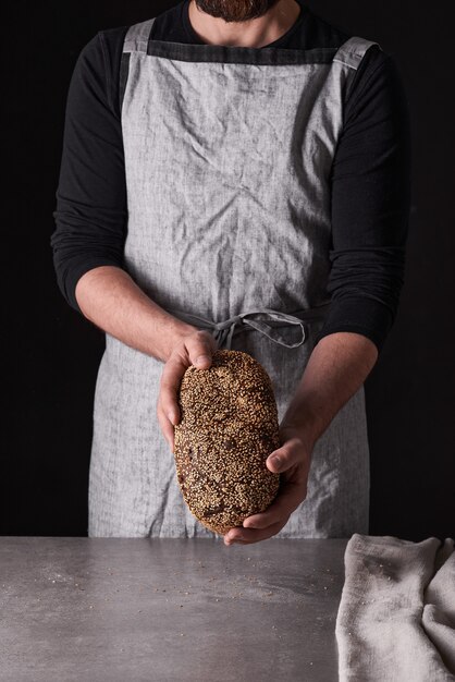 Un panadero de hombre con barba en un delantal gris está parado sobre un fondo negro y sostiene, rompe, corta pan delicioso, crujiente, panecillos, baguette.