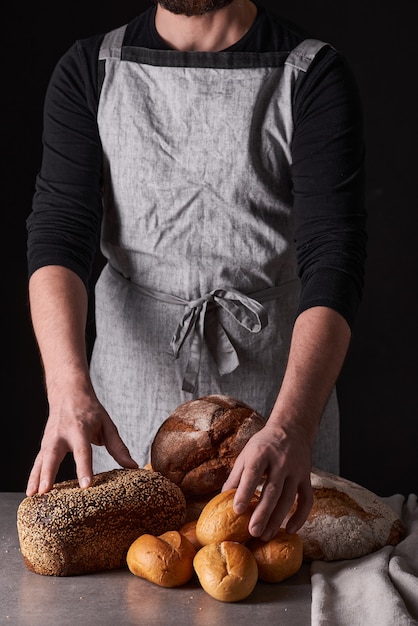 Un panadero de hombre con barba en un delantal gris está parado sobre un fondo negro y sostiene, rompe, corta pan delicioso, crujiente, panecillos, baguette.