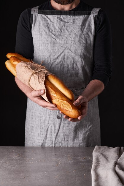 Un panadero de hombre con barba en un delantal gris está parado sobre un fondo negro y sostiene, rompe, corta pan delicioso, crujiente, panecillos, baguette.