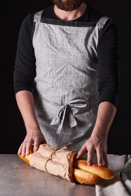 Un panadero de hombre con barba en un delantal gris está parado sobre un fondo negro y sostiene, rompe, corta pan delicioso, crujiente, panecillos, baguette.