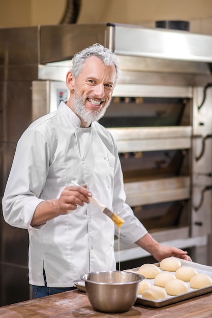 Foto panadero. hombre contento en uniforme con cepillo y cuenco cerca de bandeja de bollo de pie cocinando en panadería