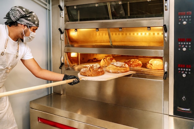 Panadero latino poniendo pan en el horno con guantes y una máscara