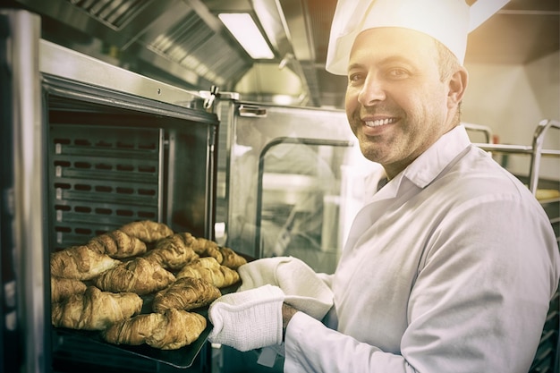 Panadero maduro sonriendo con orgullo a la cámara sacar algunos croissants del horno