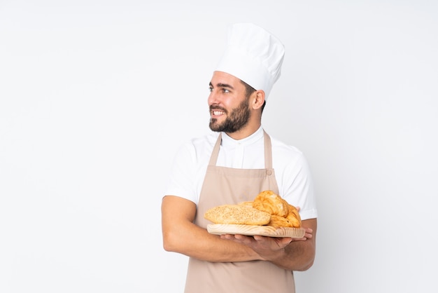 Panadero masculino sosteniendo una mesa con varios panes aislados en blanco mirando hacia el lado