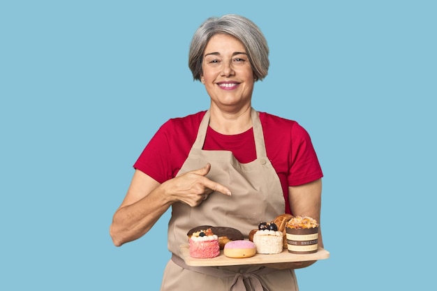 Panadero de mediana edad caucásico con bandeja de pastelería sonriendo y señalando a un lado mostrando algo en el espacio en blanco