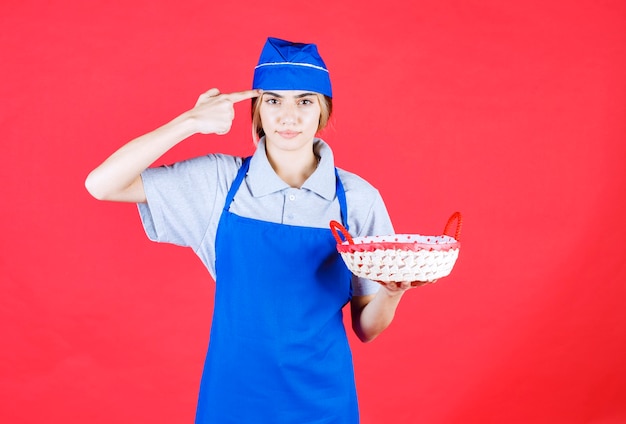 Panadero mujer en delantal azul sosteniendo una canasta de pan con una toalla roja en el interior y parece confundida y pensativa