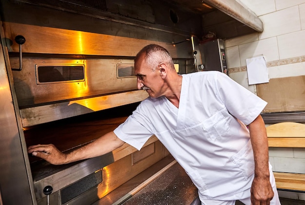 Panadero profesional en carro de inserciones uniformes con cubiertas para hornear masa cruda para hacer pan en un horno industrial en una panadería