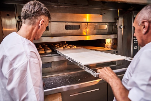 Panadero profesional en carro de inserciones uniformes con cubiertas para hornear masa cruda para hacer pan en un horno industrial en una panadería