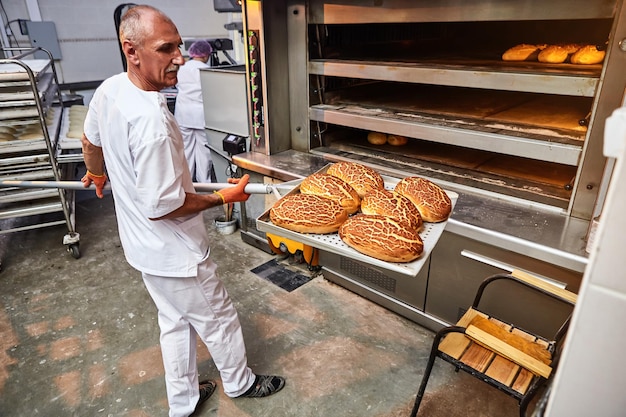 Panadero profesional en uniforme saca un carro con pan recién horneado de un horno industrial en una panadería