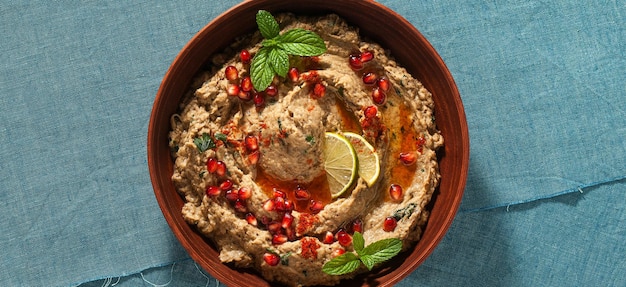 Pancarta casera de berenjena horneada baba ganoush en un bol con semillas de granada, aceite de oliva y rodajas de lima sobre un mantel de lino azul