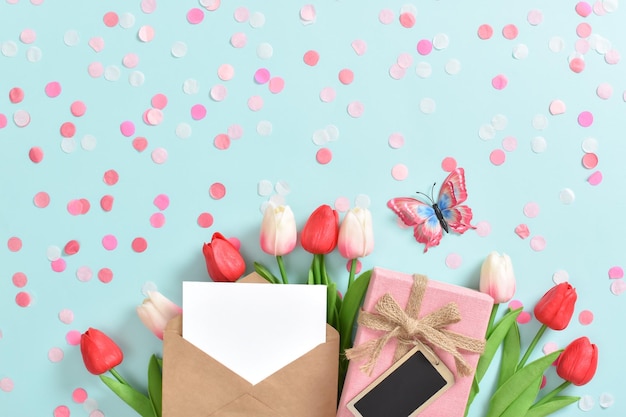 Una pancarta festiva con tulipanes y un sobre y un regalo sobre un fondo azul claro con confeti rosa Día de la Madre Día de la Mujer feliz cumpleaños Espacio de copia Vista superior plana