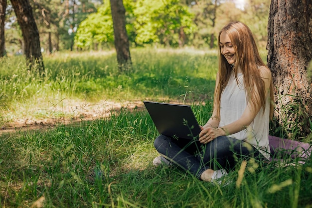 En la pancarta, una niña trabaja con una computadora portátil al aire libre en el parque sentada en el césped El concepto de trabajo remoto Trabajar como autónomo La niña toma cursos en una computadora portátil y sonríe