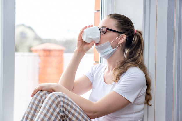 La pandemia de coronavirus. Cuarentena en casa. La mujer caucásica en baja por enfermedad está sentada en la ventana con una máscara protectora médica, bebiendo té medicinal.