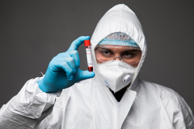 Pandemia de coronavirus. La epidemia del hombre virólogo del coronavirus chino con traje de protección, guantes, máscara y gafas examina el virus en un tubo aislado en un espacio gris.