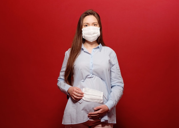 Pandemia de coronavirus, joven mujer embarazada sobre fondo rojo en máscara médica protectora sostiene el estómago