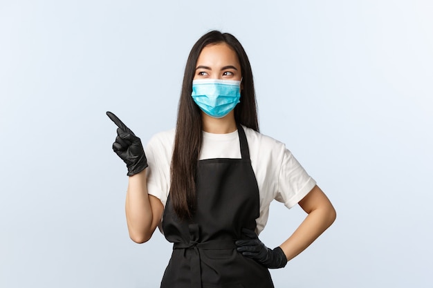 Pandemia Covid-19, distanciamiento social, pequeña empresa y concepto de prevención de virus. Sonriente mujer asiática barista, dueño de la tienda de café en máscara médica y guantes apuntando con el dedo a la izquierda en la bandera.