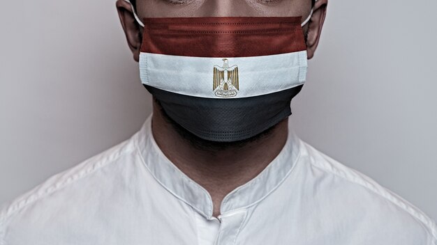 Pandemia do coronavírus. Conceito de quarentena do vírus Corona, Covid-19. O rosto masculino é coberto por uma máscara médica protetora, pintada com as cores da bandeira do Egito