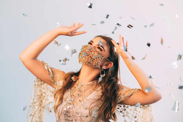 Pandemie-Party. Festliche Unterhaltung. Quarantäne-Zubehör. Glückliche Frau in kreativem Design Goldkette Gesichtsmaske passendes funkelndes Kleid genießt das Tanzen im Konfettiregen auf rosa blauem Hintergrund.