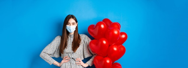 Pandemie und Valentinstag fröhliches lächelndes Mädchen in medizinischer Maske, das neben romantischem Herzballo steht