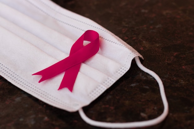 Pandemiemaske mit rosa Schleife Brustkrebsprävention auf schwarzem strukturiertem Tisch. Konzeptbild des Monats zur Prävention von Brustkrebs.