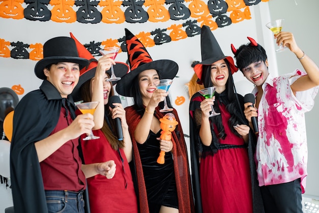 Pandilla de jóvenes asiáticos en traje de bruja, mago con celebrar la fiesta de Halloween para cantar una canción y beber, postre en la habitación. Grupo adolescente tailandés con celebrar Halloween. Concepto fiesta Halloween en casa.