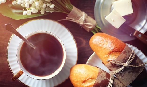 Panecillos frescos para el desayuno. Bollo con mantequilla a una taza de café por la mañana. Desayuno en el hotel, bollos, mermelada, té y ramo de flores.