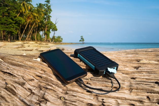 El panel solar portátil está en la playa sobre un tronco de madera y carga la batería del teléfono móvil. uso de energía solar en estado salvaje en una isla desierta. estanco, impermeable, estanco