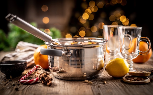 Panela de aço e ingredientes para a preparação de vinho quente na mesa de madeira em frente à iluminação festiva