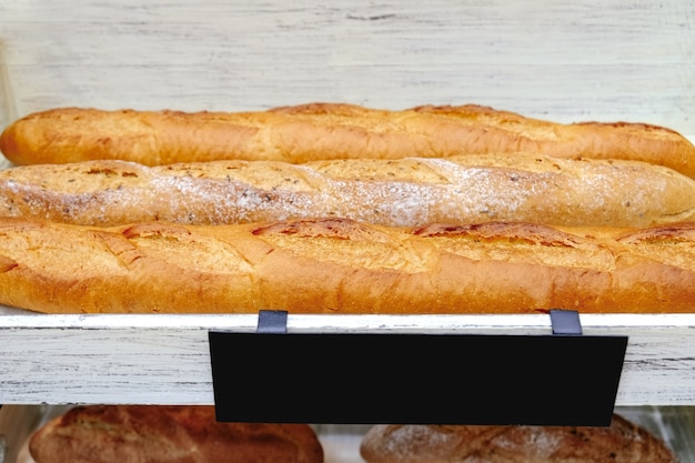 Panes baguette sin gluten recién horneados en estantes de madera blancos con una etiqueta de etiqueta negra en blanco.