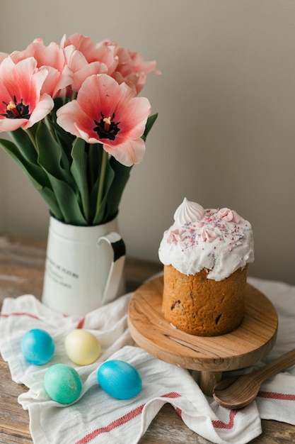 Panettone de Pascua y huevos coloreados de Pascua. Símbolos de Pascua. Tulipanes rosas en jarra de metal sobre mesa de madera