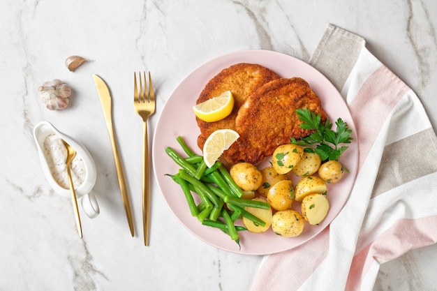 Paniertes Schweineschnitzel mit jungen Kartoffeln, grünen Bohnen mit Besteck und Mayonnaise-Soße serviert auf einem rosa Teller mit Zitronenspalten, auf einer hellen Marmoroberfläche, Draufsicht, Nahaufnahme