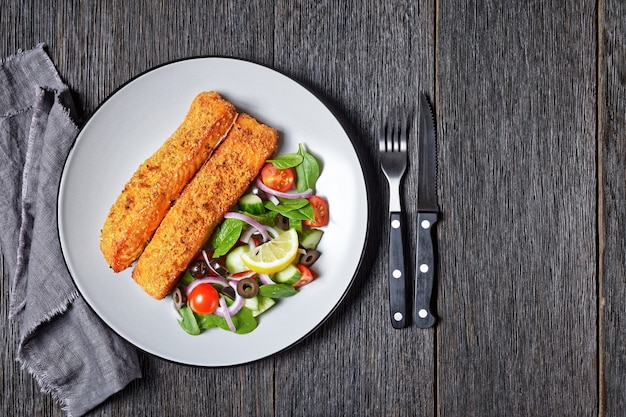 Panko verkrustete gebackene Lachsfilets mit Spinat-Tomaten-Gurken-Oliven-Salat auf einem Teller auf einem dunklen Holztisch, Ansicht von oben, flach, freier Raum