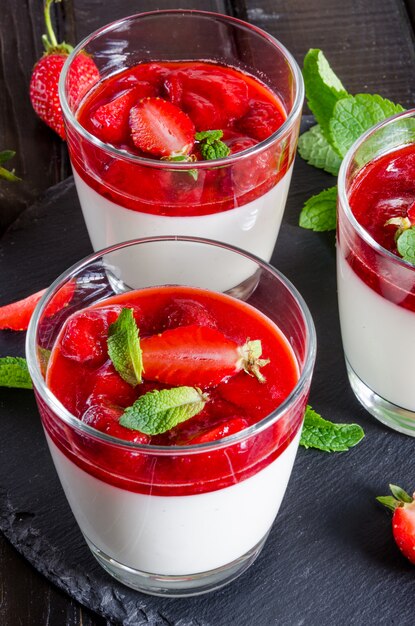 Panna cotta italiana tradicional en un vaso con salsa de fresas en una mesa de madera oscura.