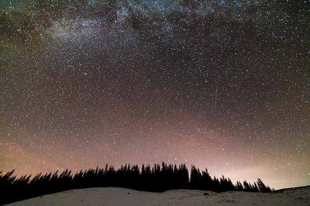 Panorama de paisagem de noite de montanhas de inverno. Constelação brilhante da Via Láctea no céu estrelado azul escuro sobre a floresta de pinheiros de abeto vermelho escuro, brilho suave no horizonte após o pôr do sol.